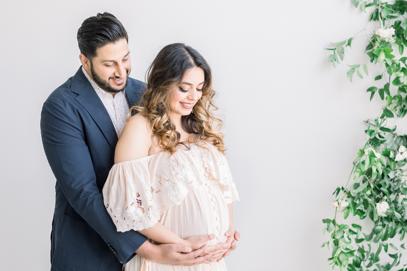 Dad hugging mom during studio maternity session with florals in background. Photo by Sana Ahmed Photography.