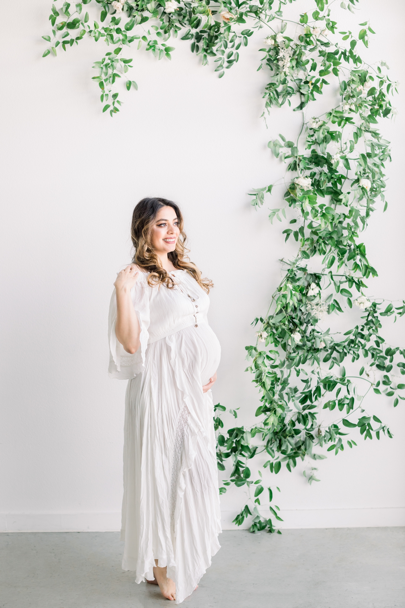 Pregnant mom wearing white maxi dress in studio with florals in the background. Photo by Sana Ahmed Photography.