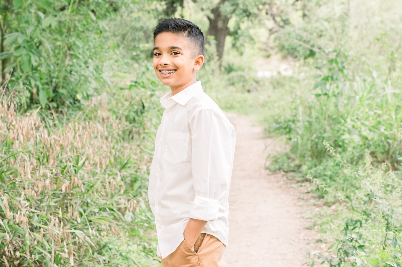 Child smiling at camera with his hands in his pockets in beautiful field. Photo by Austin family photographer, Sana Ahmed Photography.