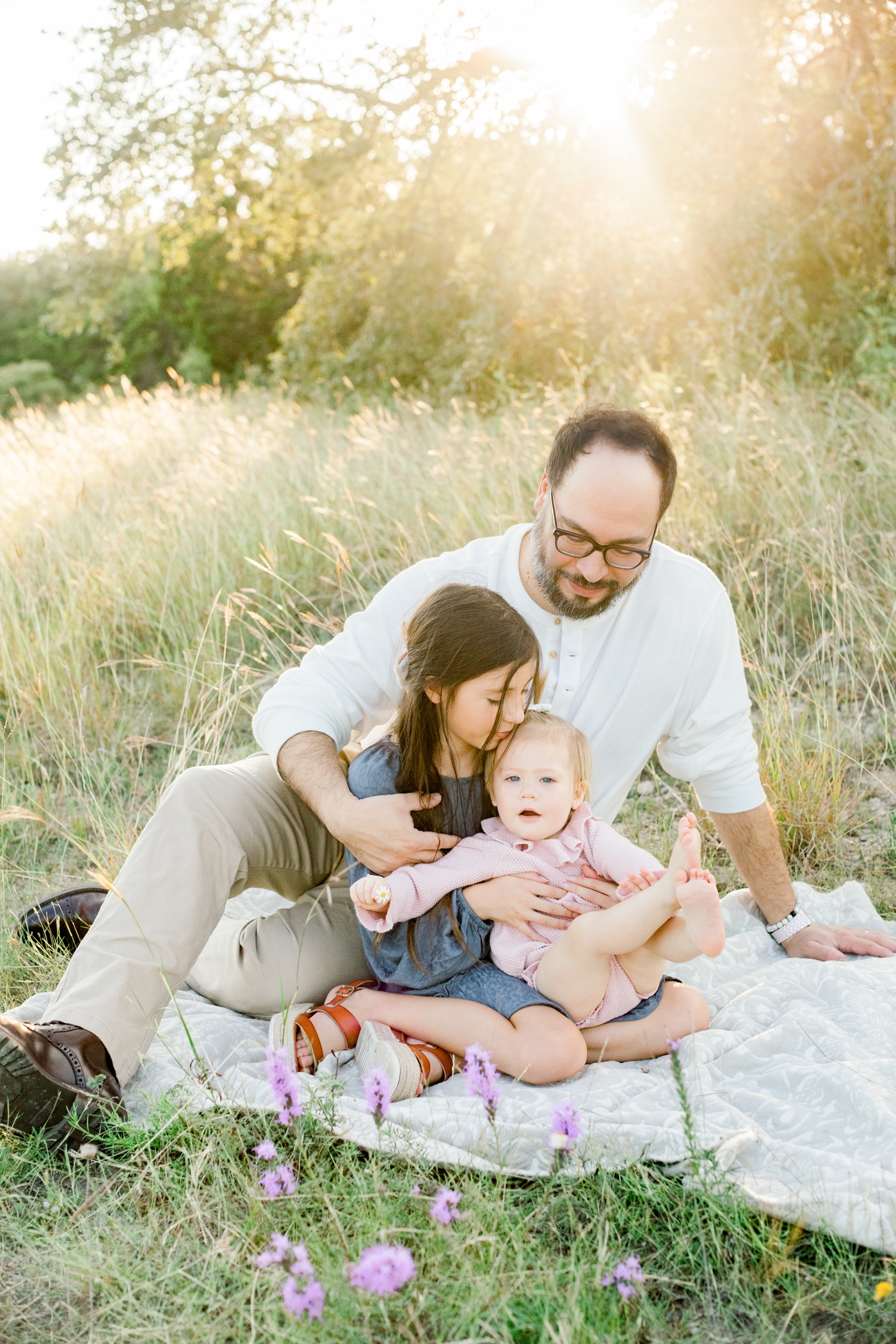 Dad with two daughters sitting on a blanket during a sunny field session. Photo by Austin Texas family photographer, Sana Ahmed Photography