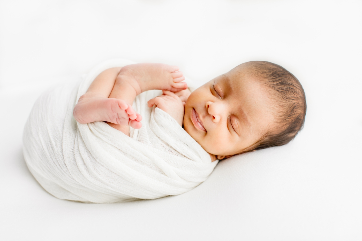 Newborn baby boy curled up in white swaddle on white backdrop during Austin TX newborn session. Photo by Sana Ahmed Photography.