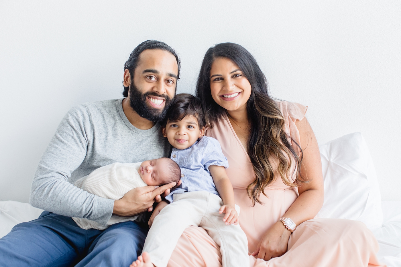 Sweet family photo of parents and toddler smiling at camera during newborn session. Photo by Sana Ahmed Photography.