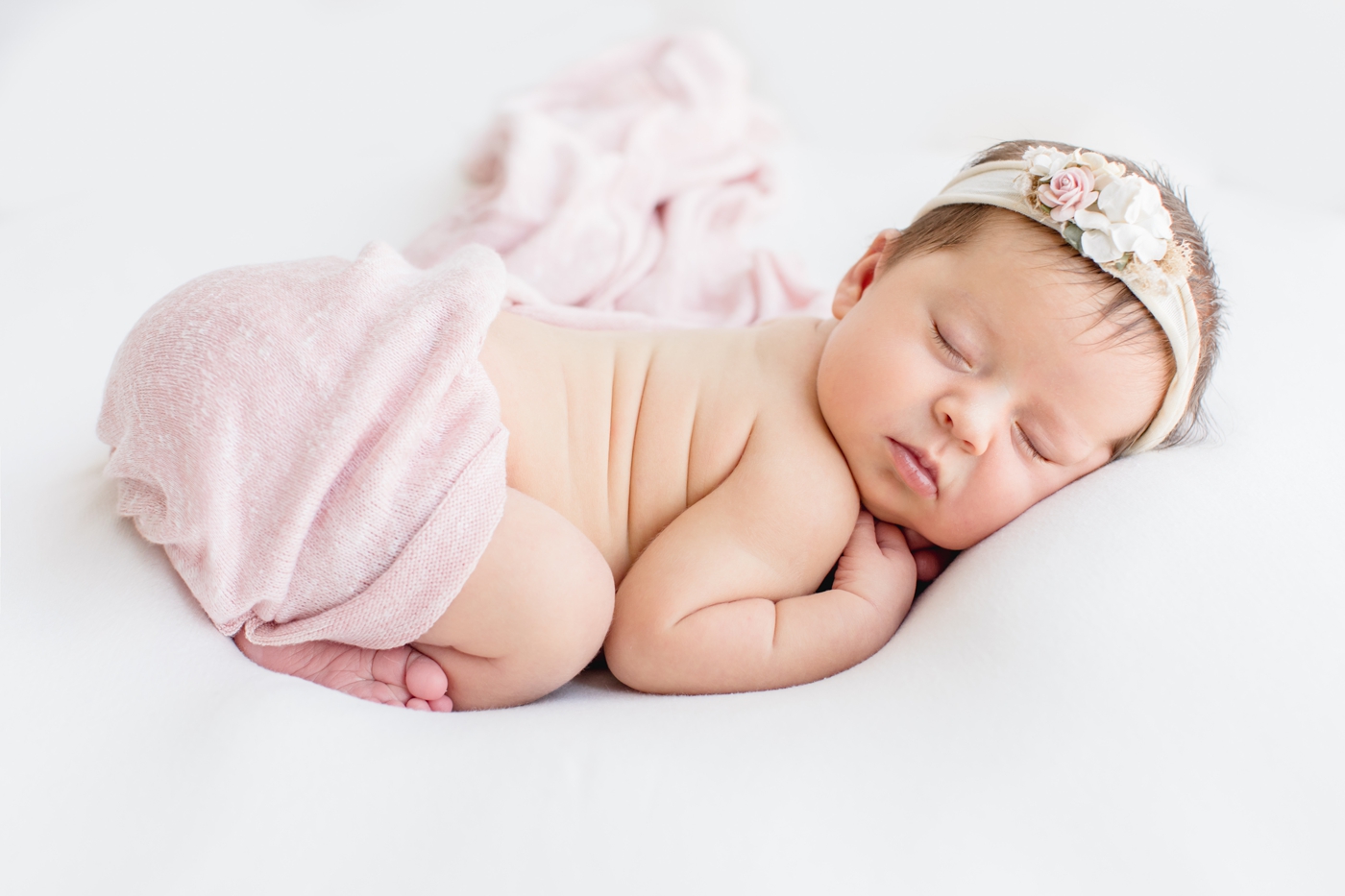 Baby girl on white background with pink swaddle and floral headband during studio newborn session in Austin TX. Photo by Sana Ahmed Photography.
