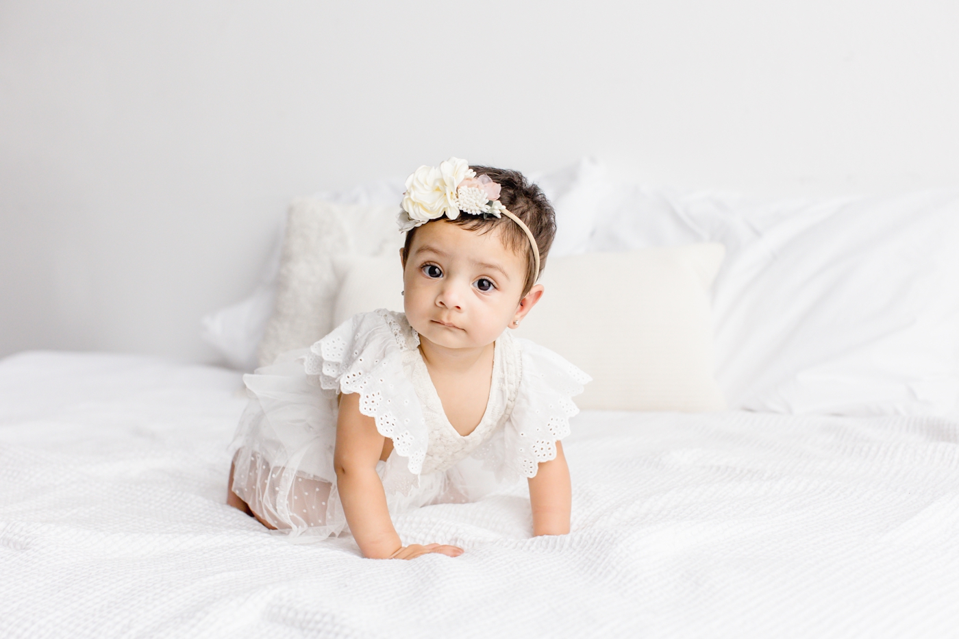 Baby girl in white dress and floral bow looking at camera during milestone session with Sana Ahmed Photography.