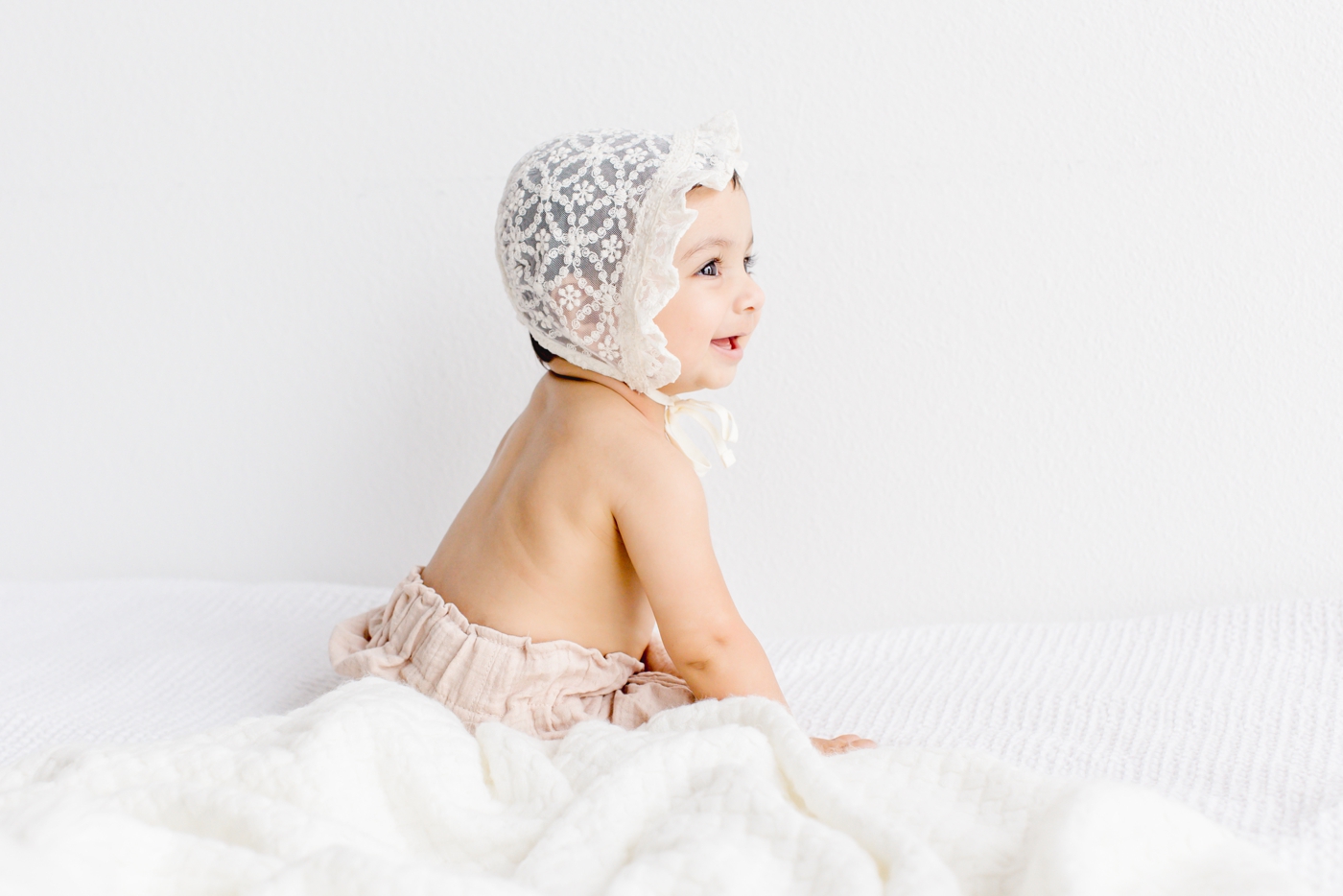 Baby girl in white lace bonnet smiling during studio session. Photo by Sana Ahmed Photography.