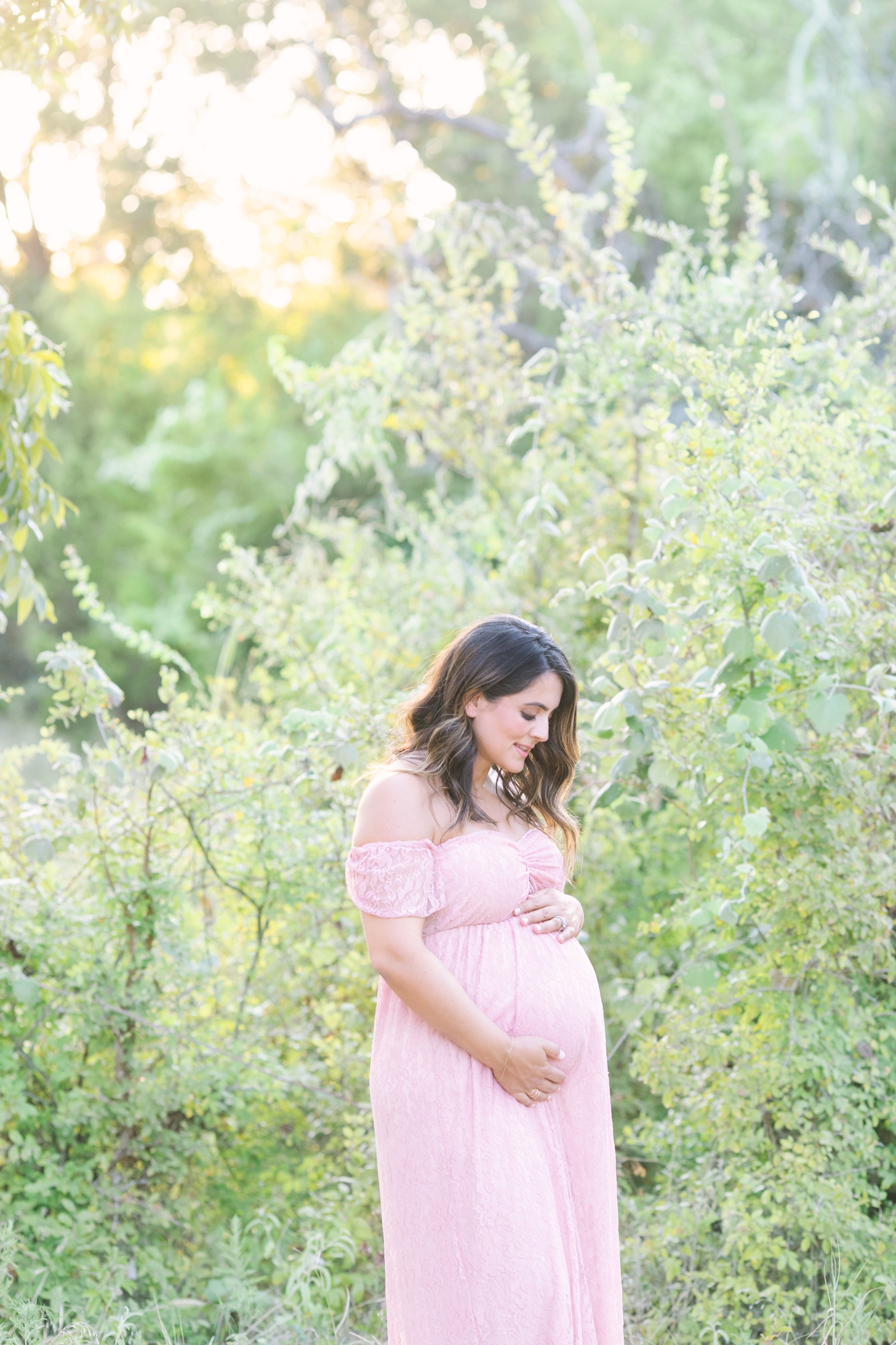 Mom wearing pink lace maxi dress during maternity session in Austin park with golden light coming through the trees. Photo by Sana Ahmed Photography.