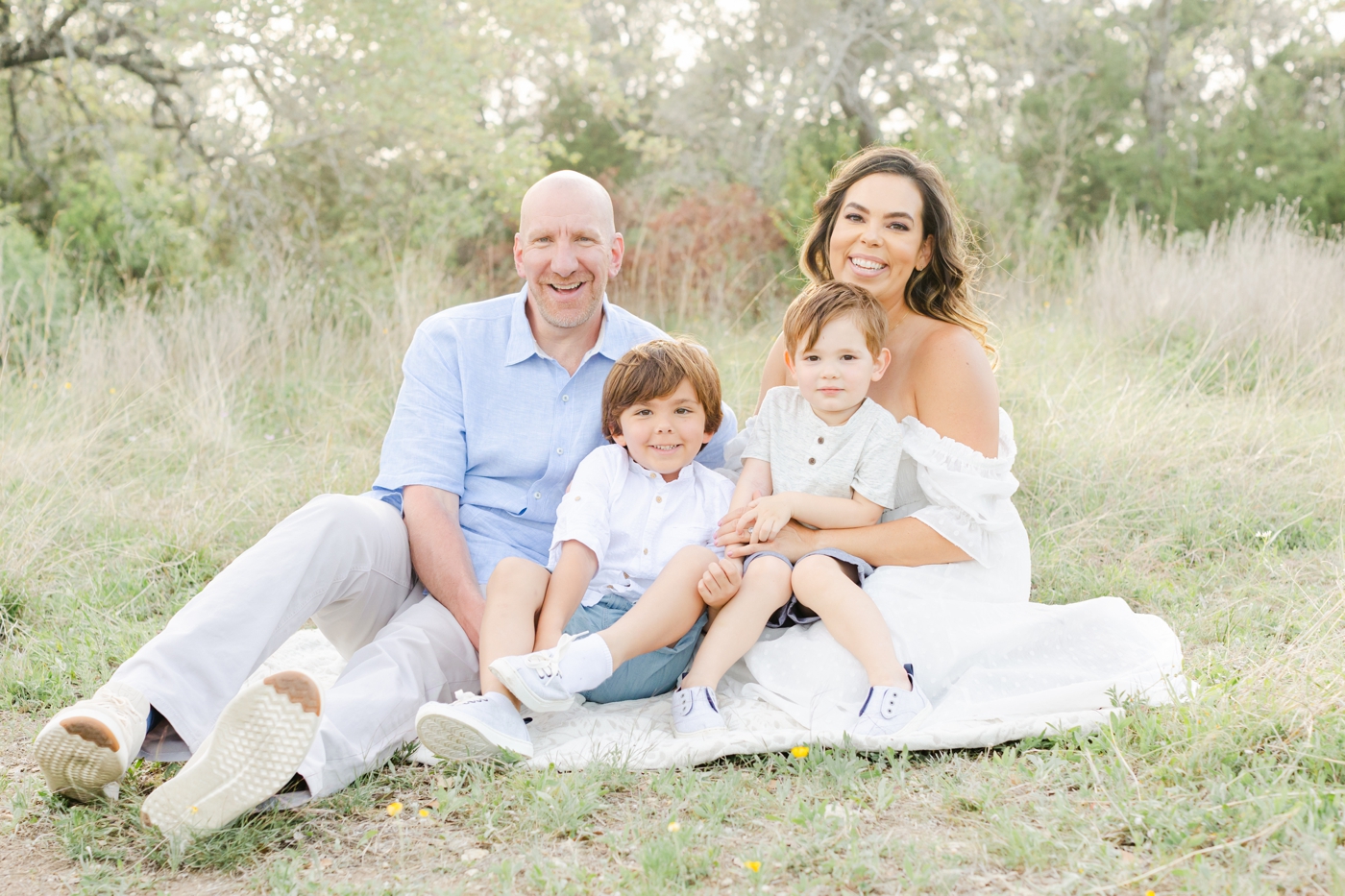 Beautiful family smiling at camera while sitting on a white blanket during photoshoot. Photo by Cedar Park family photographer, Sana Ahmed Photography.
