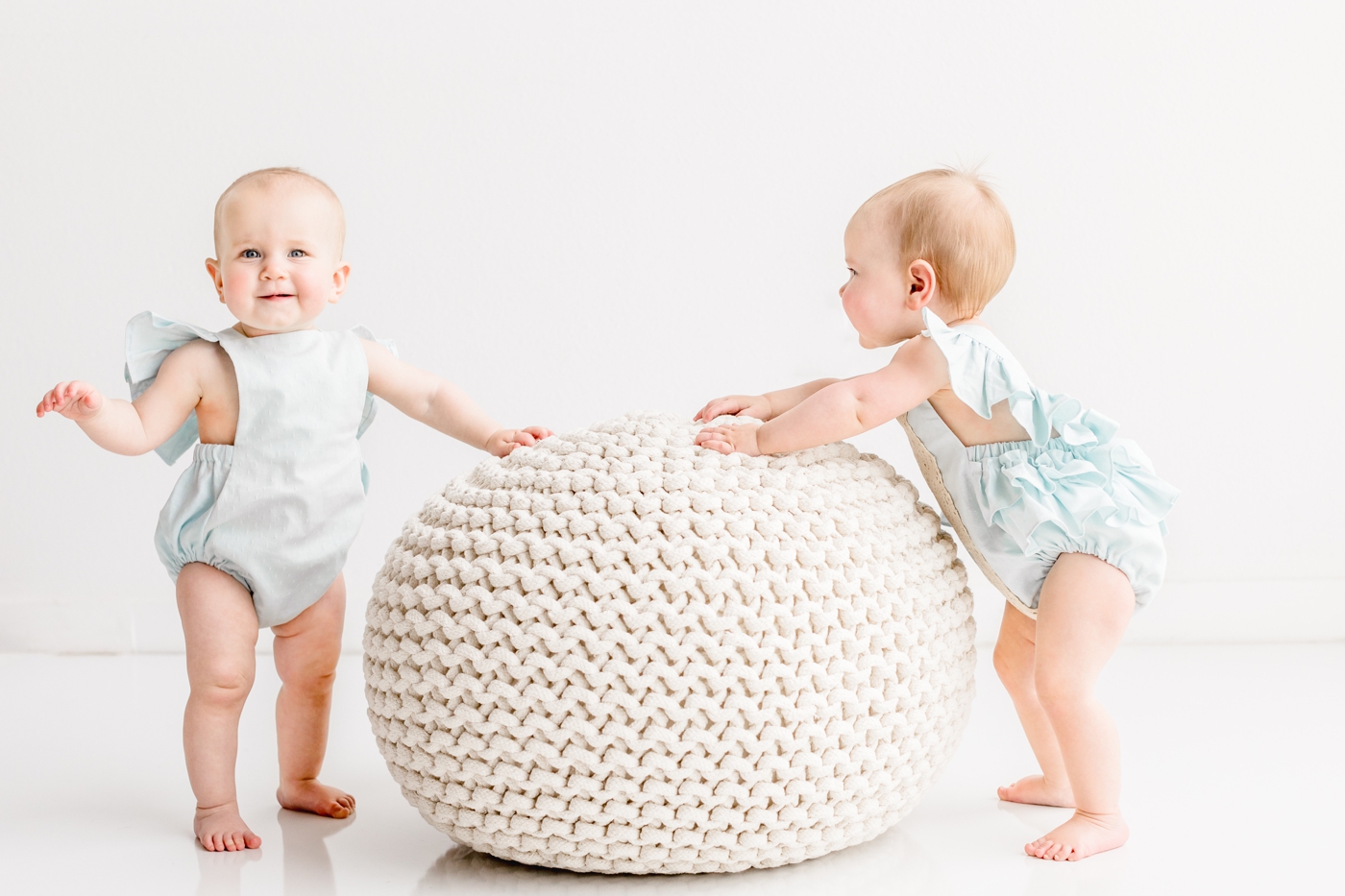 Twin girls using knit pouf to support them as they stand during milestone photo session. Photo by Sana Ahmed Photography.