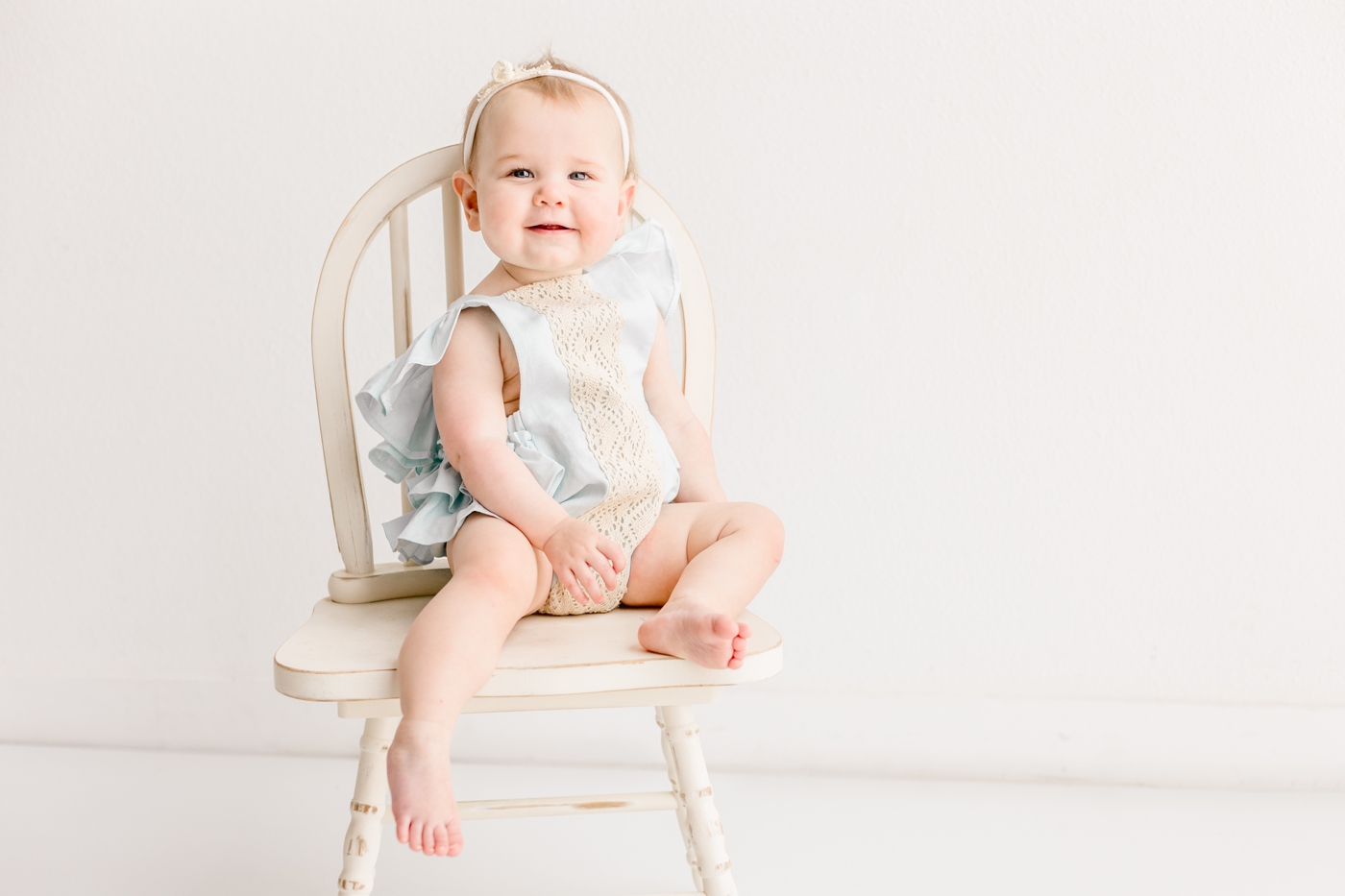 Smiling baby girl sitting on wooden chair during first birthday session in Austin, TX. Photo by Sana Ahmed Photography.