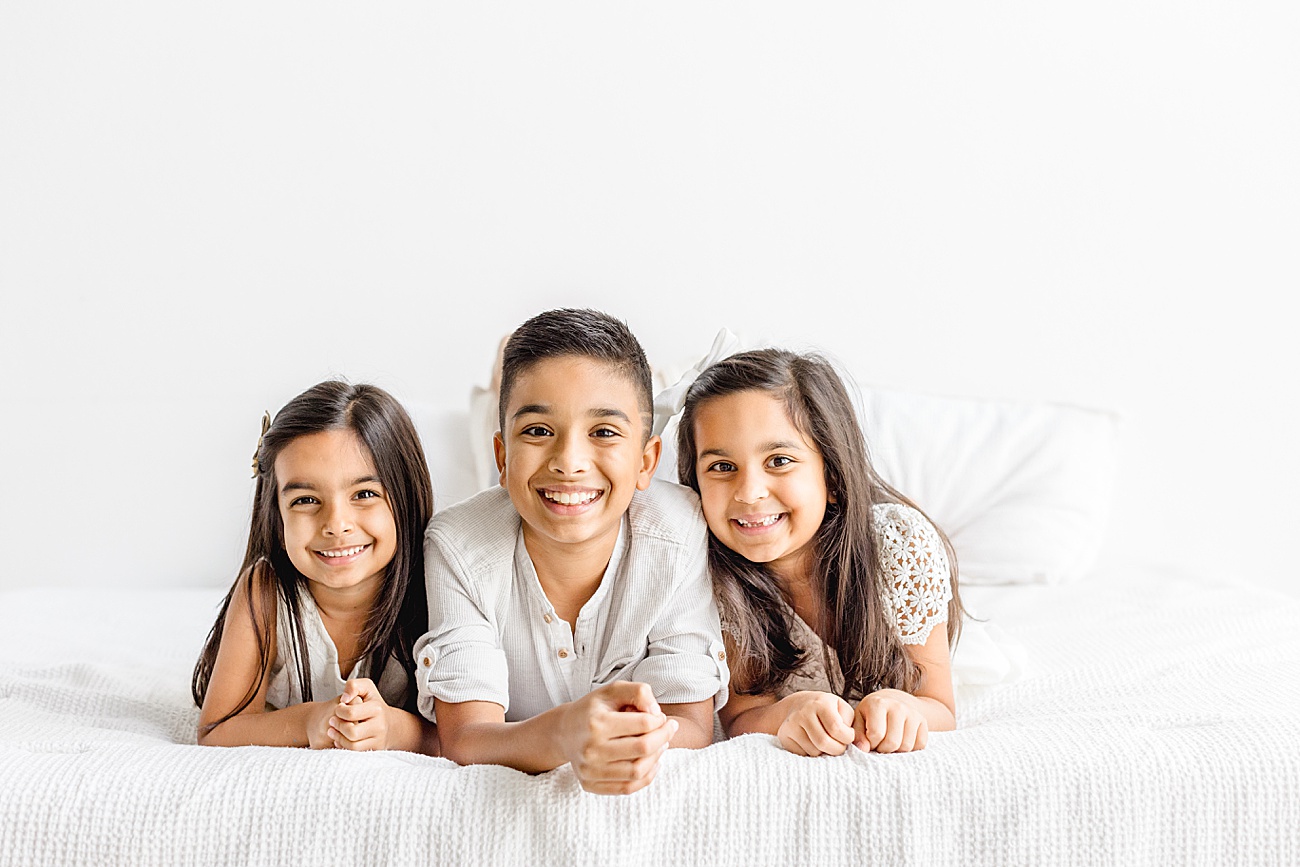Sweet portrait of three siblings smiling at camera while laying on studio bed. Photo by Sana Ahmed Photography.