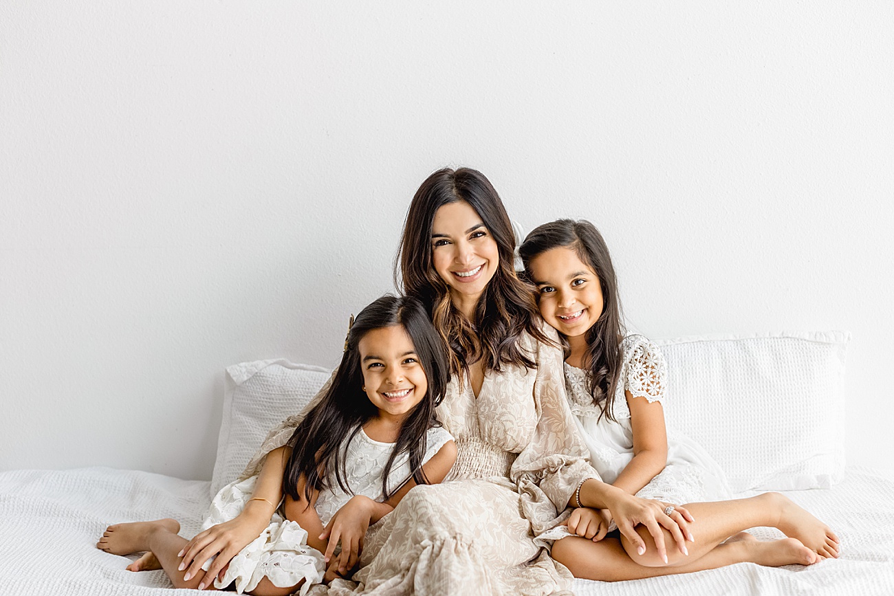 Mom sitting on bed with daughters smiling at camera. Photo by Sana Ahmed Photography.