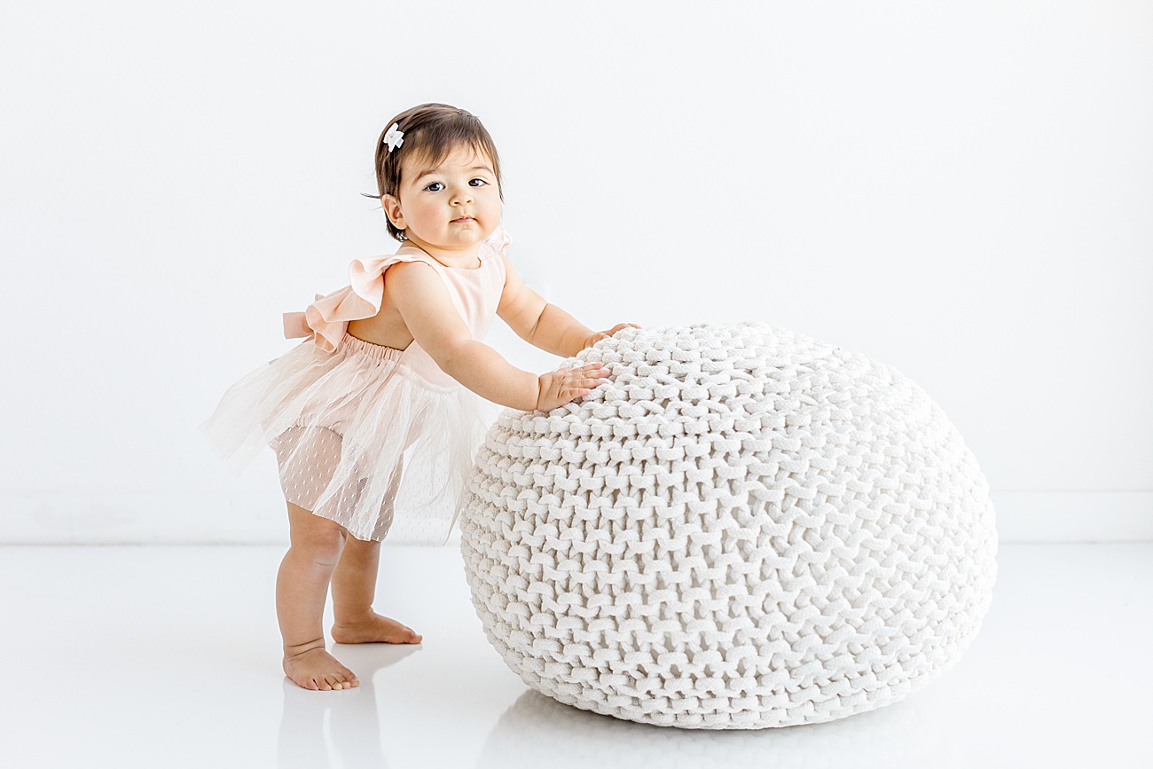 Baby holding onto white knit pouf in studio as she stands for photo with Sana Ahmed Photography