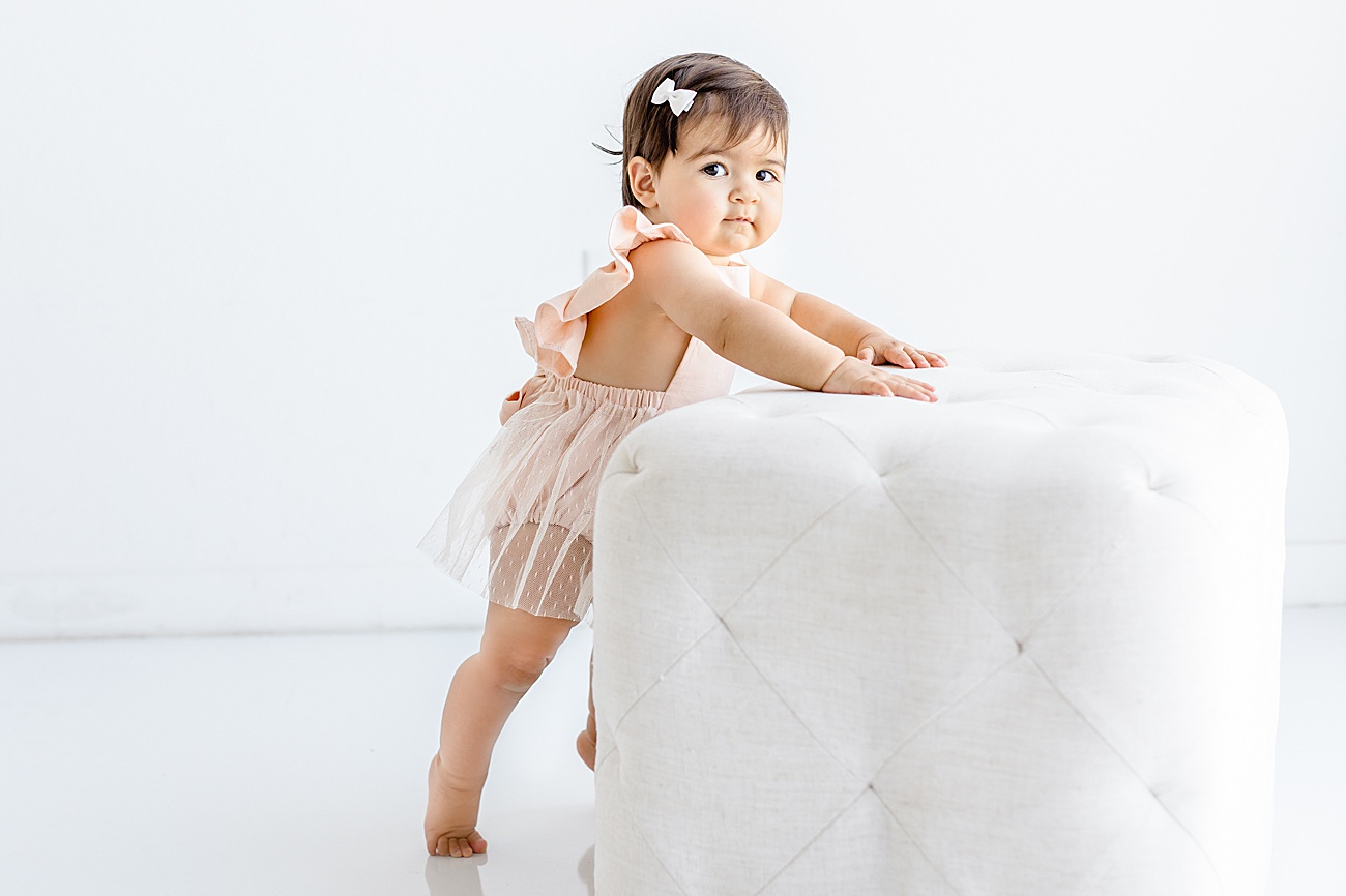 Baby leaning on white ottoman with cute pink flutter sleeve romper. Photo by Sana Ahmed Photography