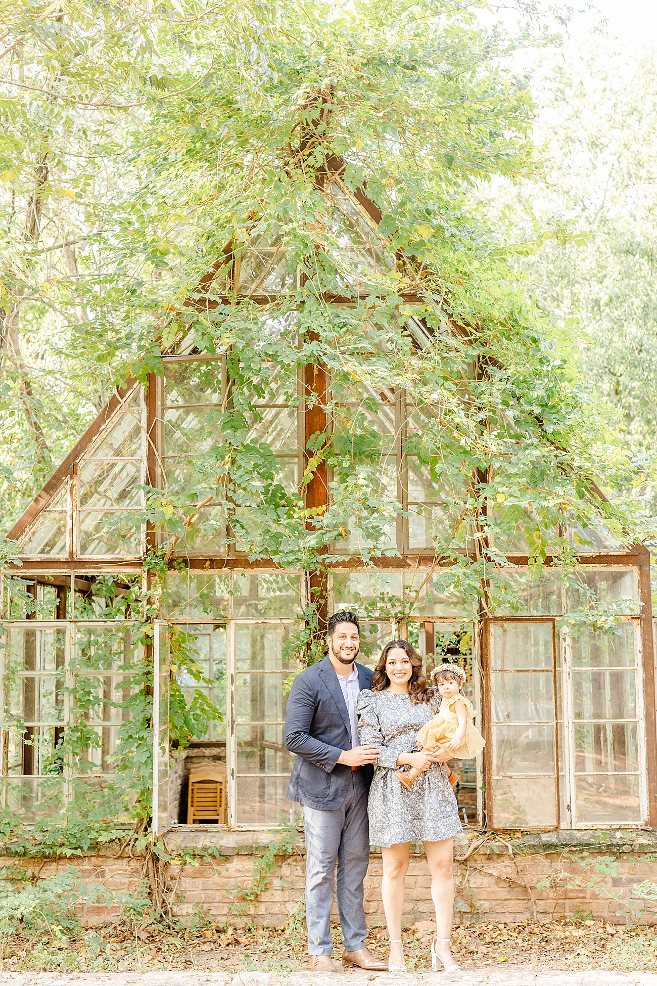 Family smiling at camera outside of vintage greenhouse at unique location for family photos in Austin, Texas. Photo by Sana Ahmed Photography