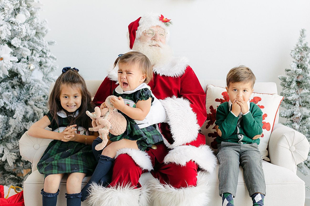 Photo of baby crying on Santa's lap while siblings react. Image by Sana Ahmed Photography.