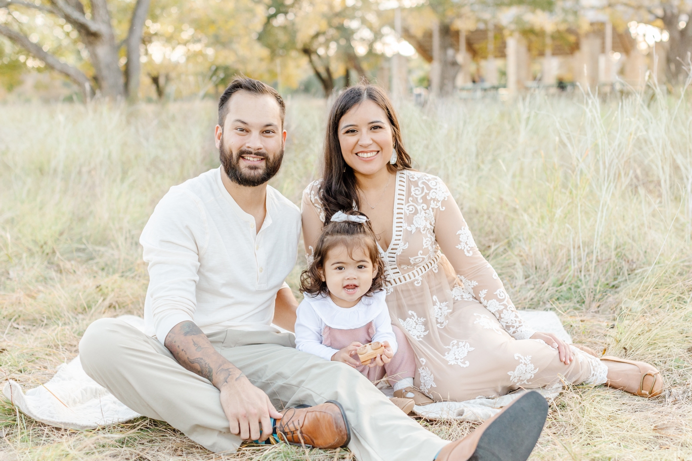 Family smiling at the camera as they snuggle on the grass during park family session in Austin. Photo by Sana Ahmed Photography.