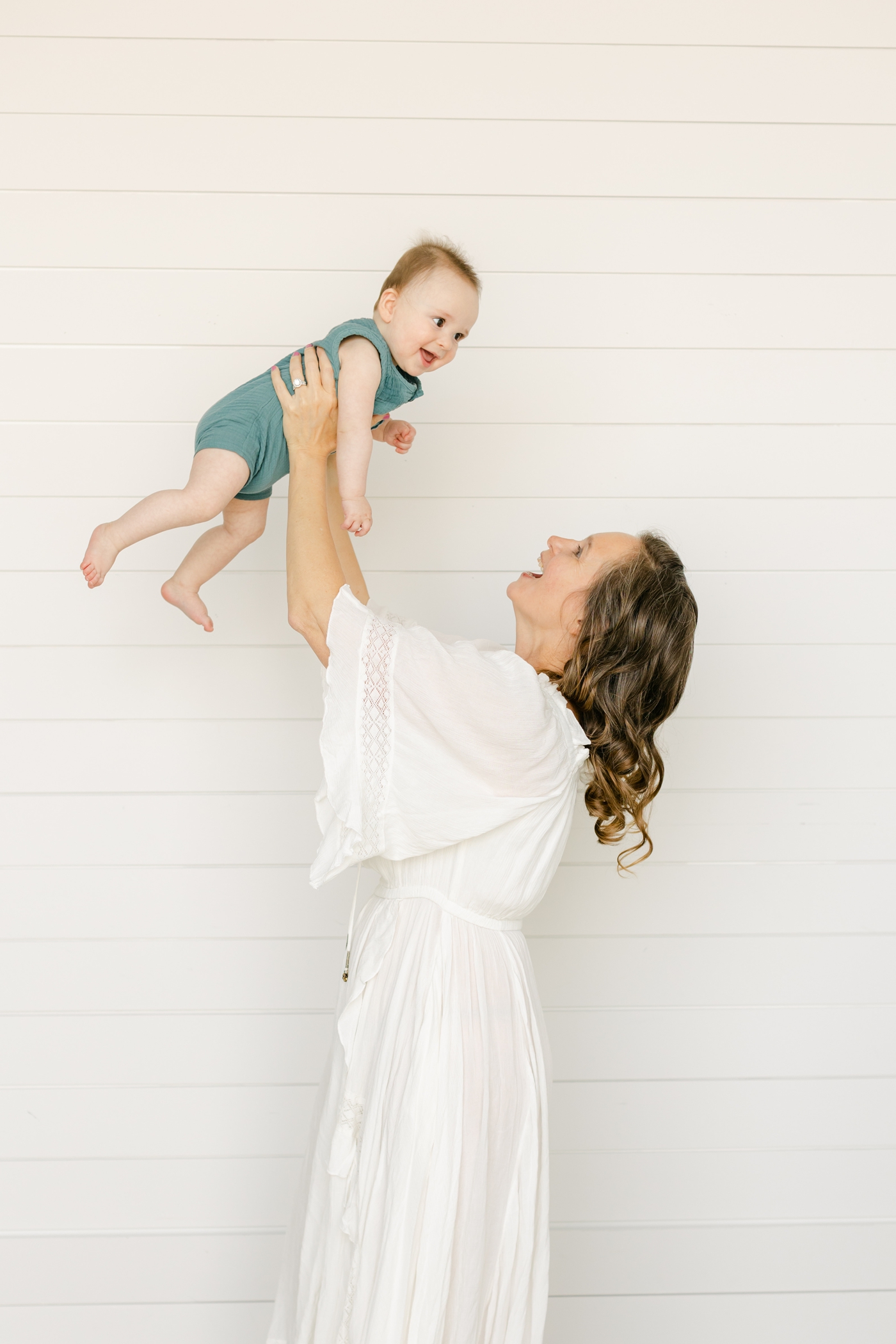 Mom playing airplane with baby in blue romper with shiplap in the background. Photo by Sana Ahmed Photography.