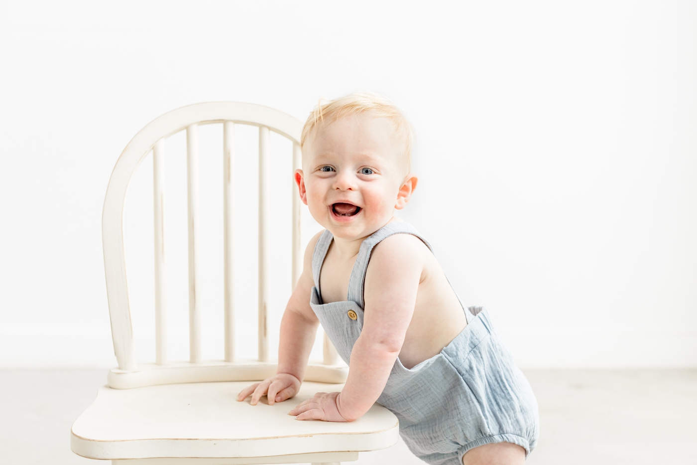 Little boy smiling at camera while leaning on white chair. Photo by Sana Ahmed Photography.