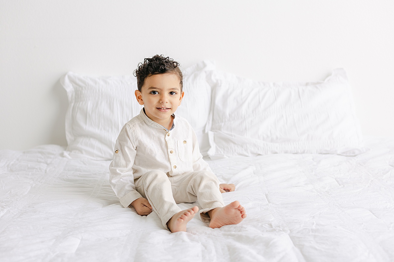 Toddler boy smiling as he sits on bed in light and airy studio in Austin, TX. Photo by Sana Ahmed Photography.