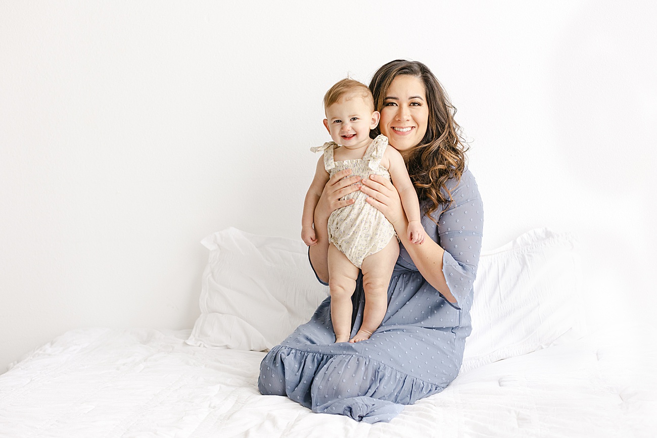 Baby and Mom smiling big at camera while sitting on studio bed in Austin, TX. Photo by Sana Ahmed Photography.