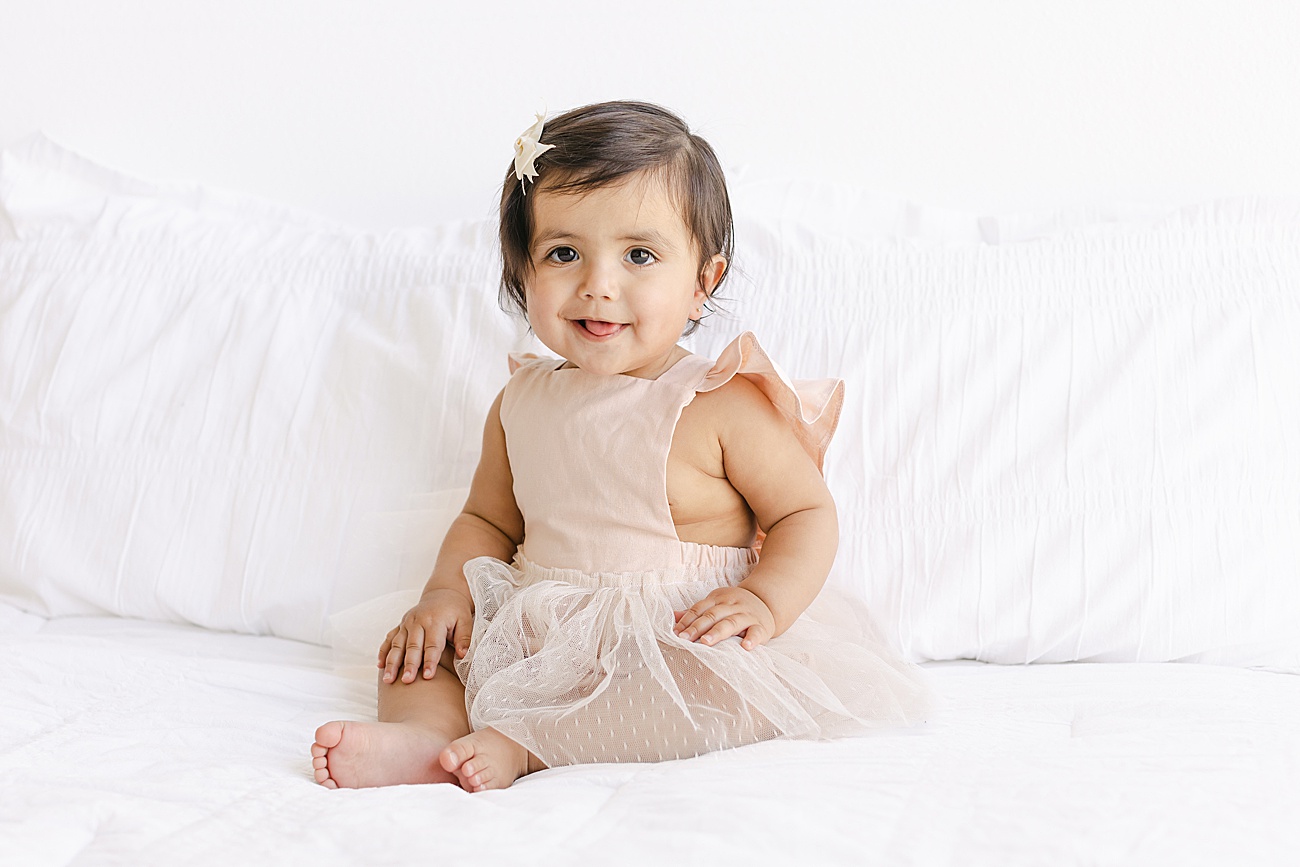 Little girl sitting on studio bed smiling at camera during milestone photos in Austin Texas. Photo by Sana Ahmed Photography.