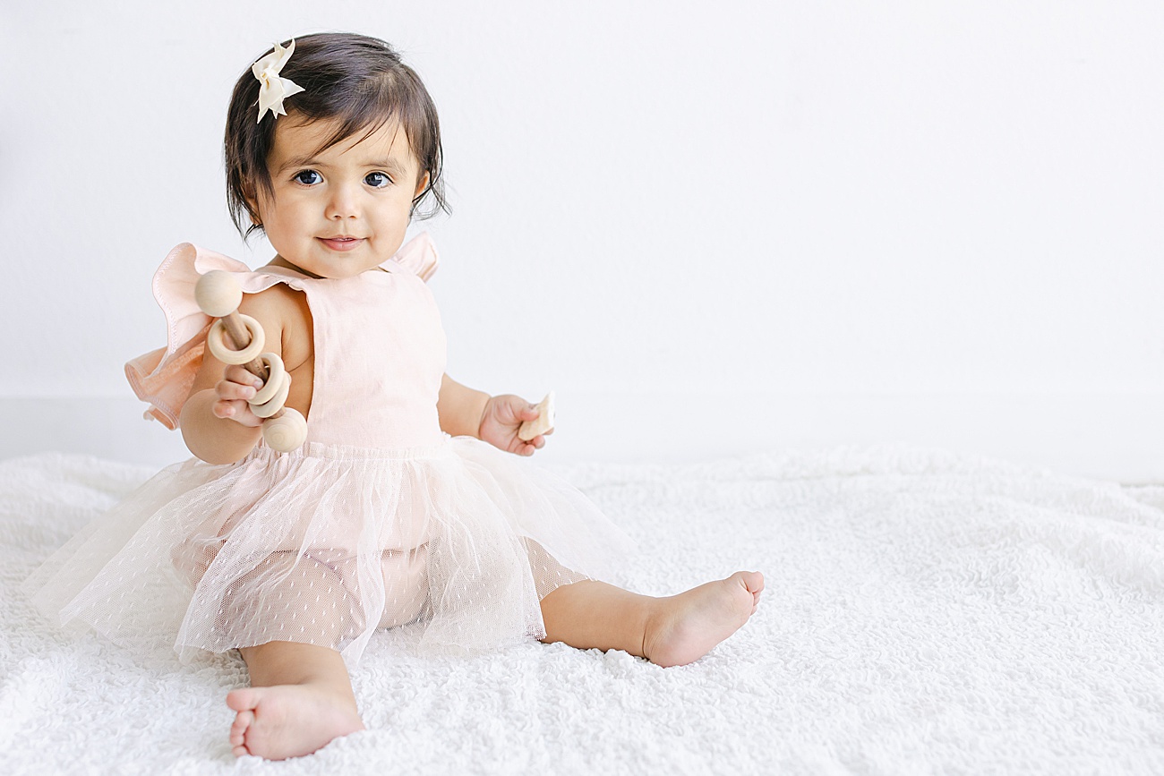Birthday girl holding wooden rattle while wearing pink romper with tulle skirt. Photo by Sana Ahmed Photography.