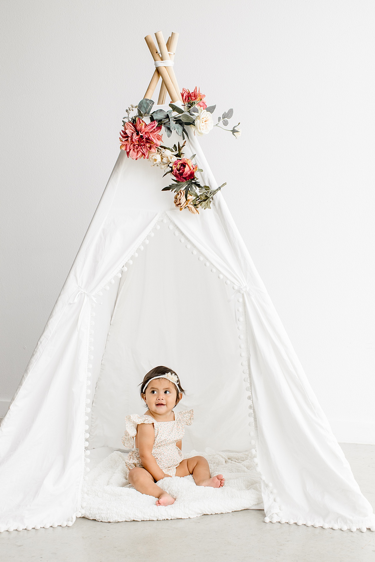 Little girl sitting in white teepee with floral decor during first birthday milestone photos in Austin Texas. Photo by Sana Ahmed Photography.