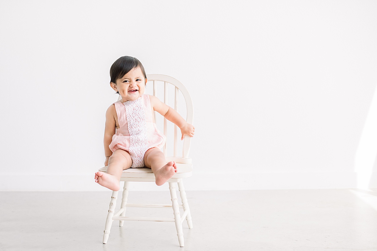 Little girl sitting on chair smiling in natural light studio. Photo by Sana Ahmed Photography.