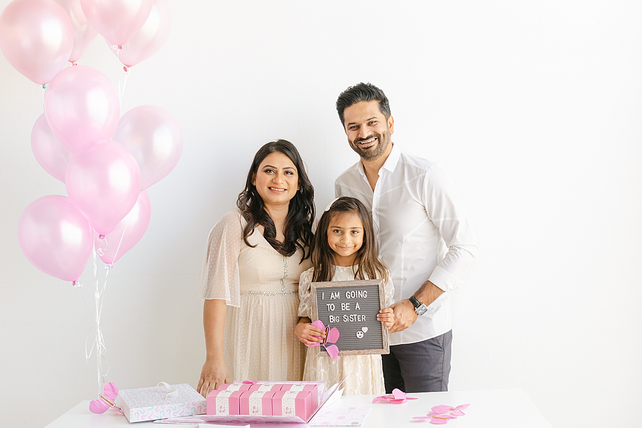 Family smiling at the camera as they hold a letter board and pink paper from their gender reveal session.Photo by Sana Ahmed Photography.