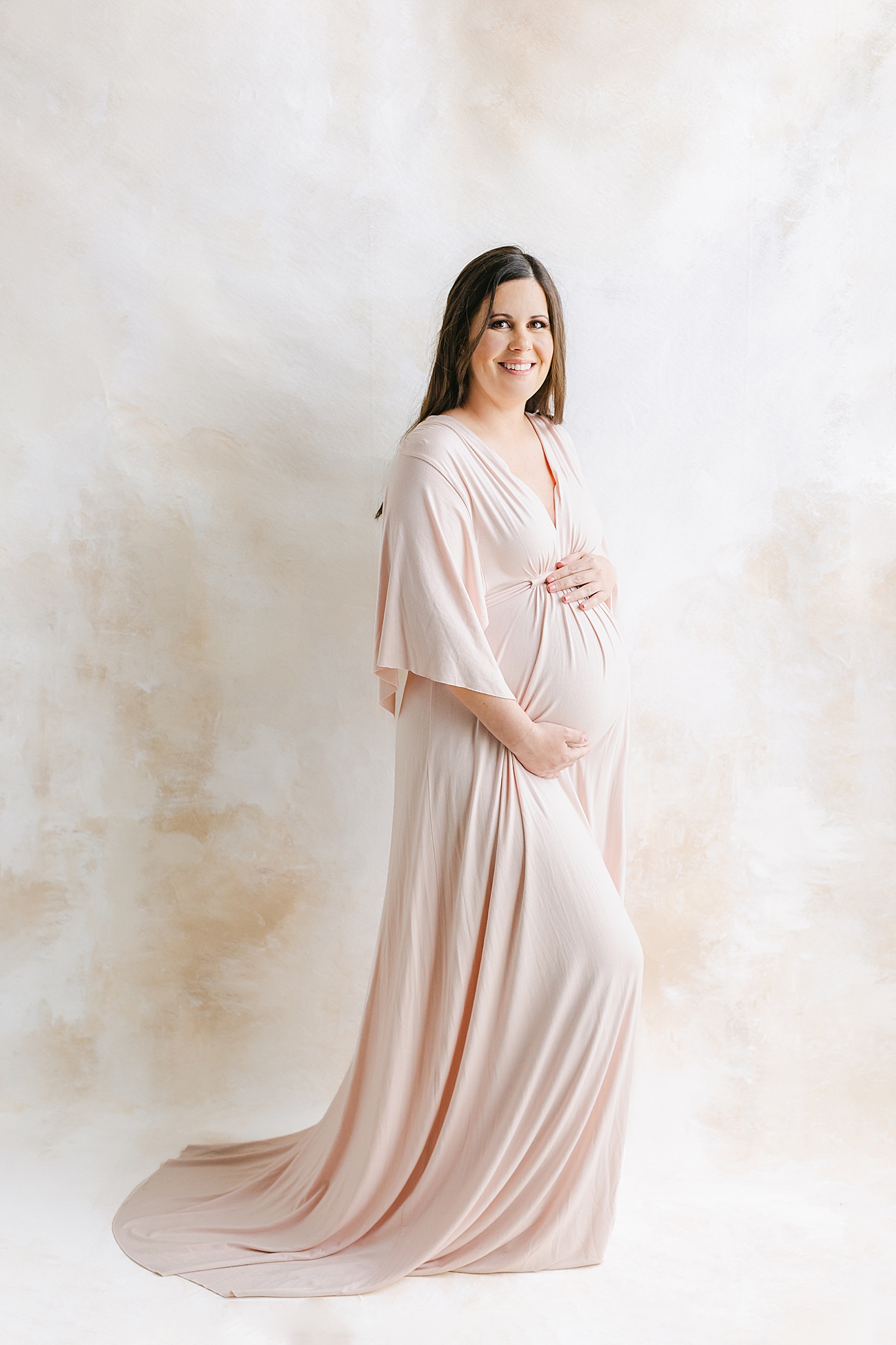 Expecting Mom wearing blush maxi dress in front of custom, hand-painted backdrop during maternity photos in Austin, TX studio. Photo by Sana Ahmed Photography.