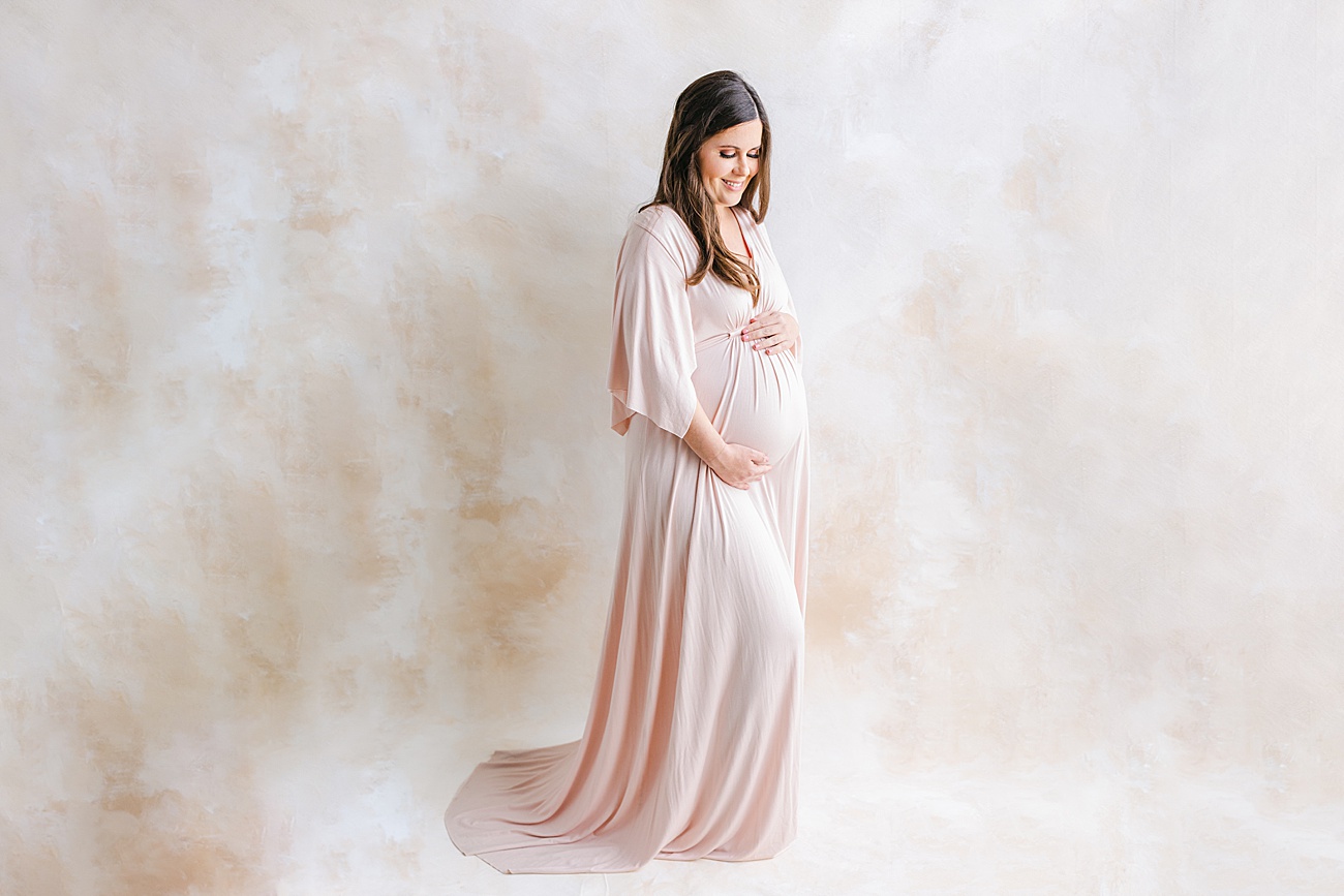 Stunning Mama looking down at baby bump in front of pastel-colored hand-painted backdrop. Photo by Sana Ahmed Photography.