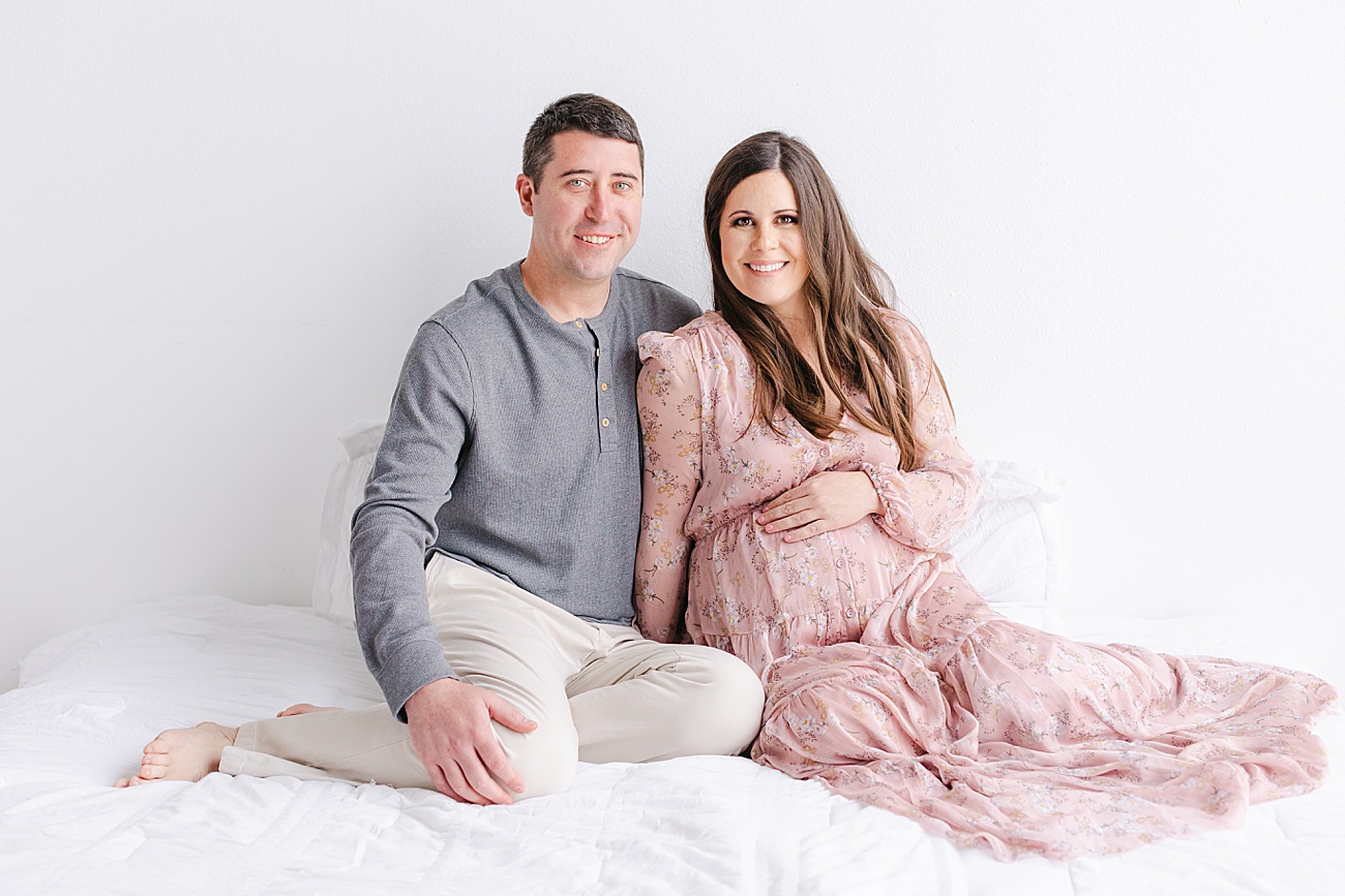 Maternity photo of Mom and Dad smiling at camera while sitting on bed in Austin, TX studio. Photo by Sana Ahmed Photography.