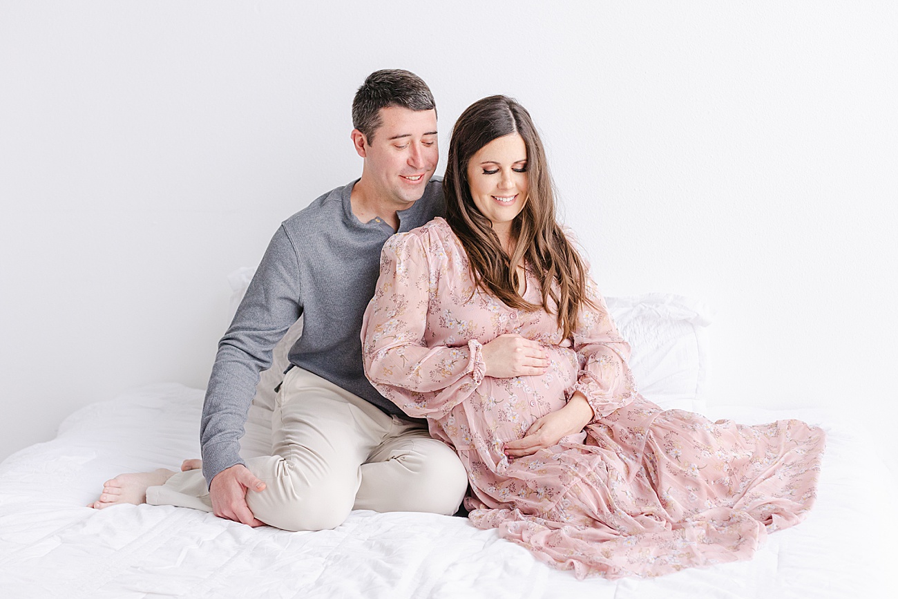 Couple looking at baby bump during maternity photos as they sit on bed in studio. Photo by Sana Ahmed Photography.