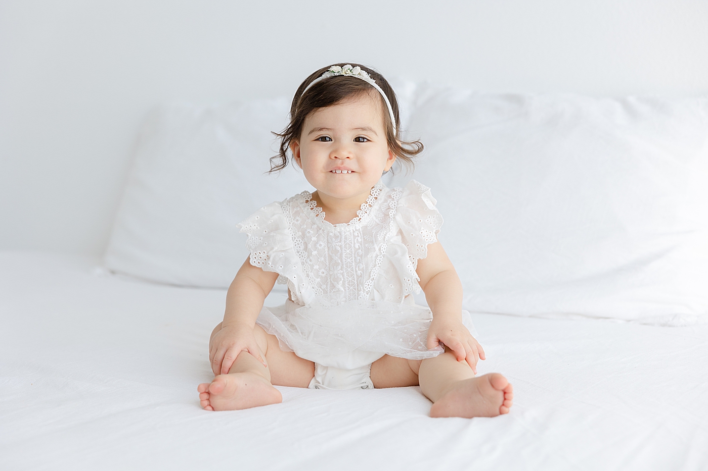 Baby girl in a white dress and headband smiling while sitting on a bed | Image by Sana Ahmed Photography