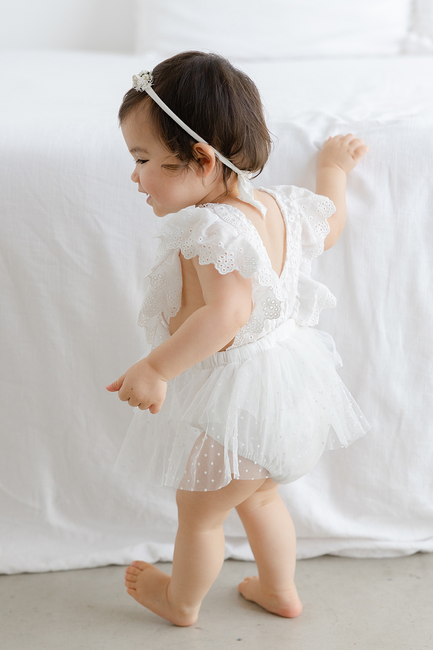 Baby girl in a white dress and headband standing | Image by Sana Ahmed Photography