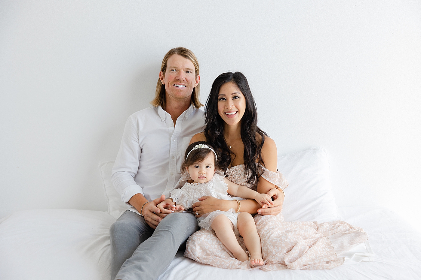 Mom dad and baby girl smiling sitting together on a bed | Image by Sana Ahmed Photography
