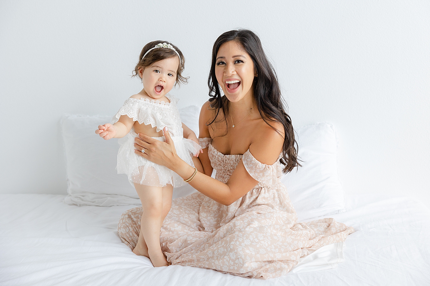 Mom and baby girl sitting on a white bed during her one year studio milestone session | Image by Sana Ahmed Photography