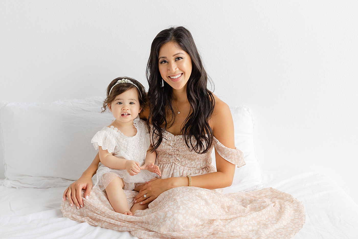 Mom and baby girl sitting on a white bed during her one year studio milestone session | Image by Sana Ahmed Photography
