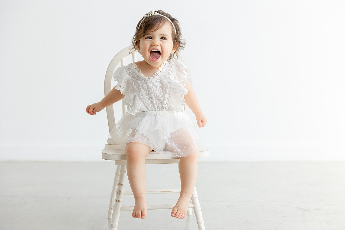 Baby girl sitting on a white chair during her one year studio milestone session | Image by Sana Ahmed Photography
