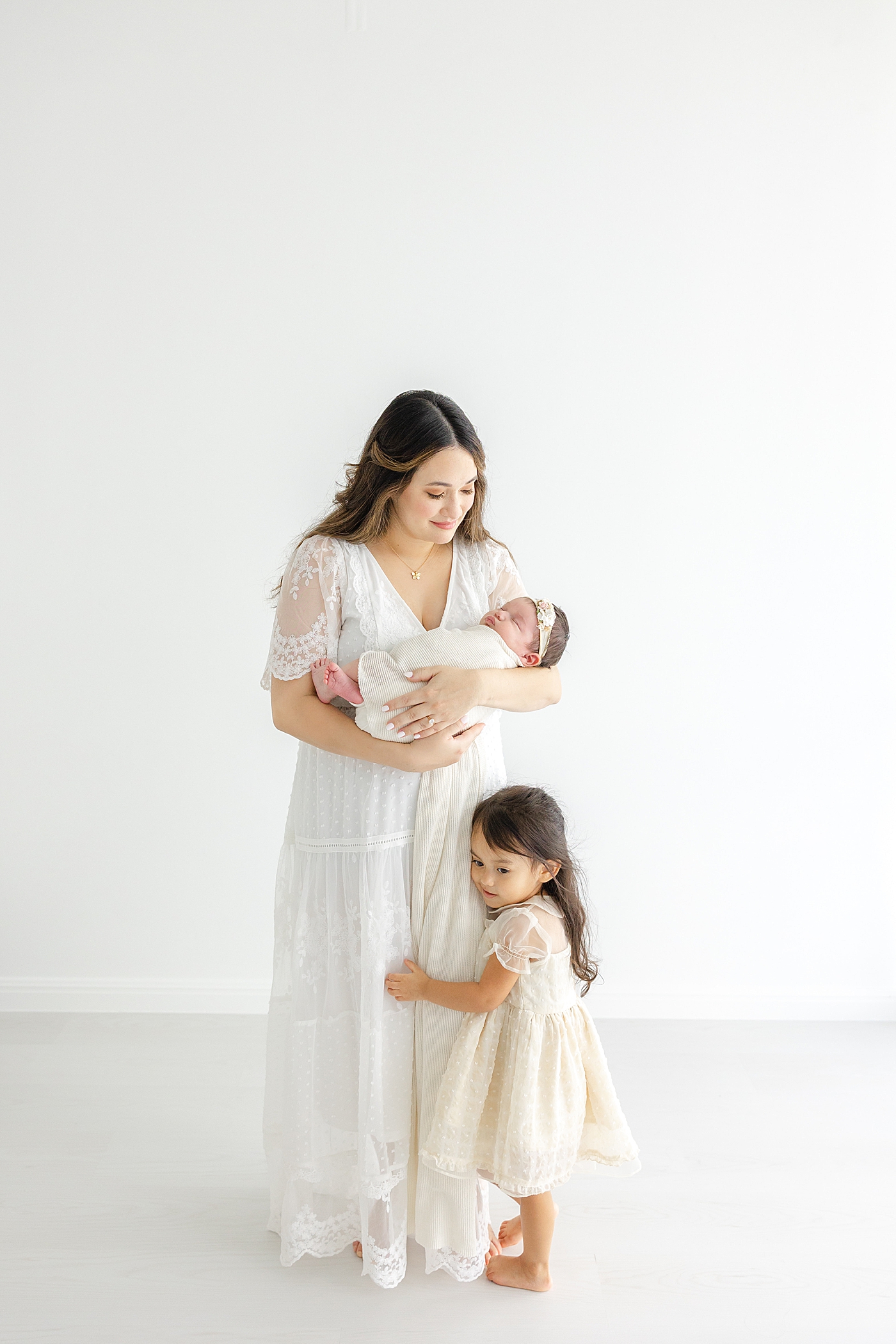 Mom in white holding her new baby girl with daughter during her Austin Studio Newborn Session | Photo by Sana Ahmed Photography