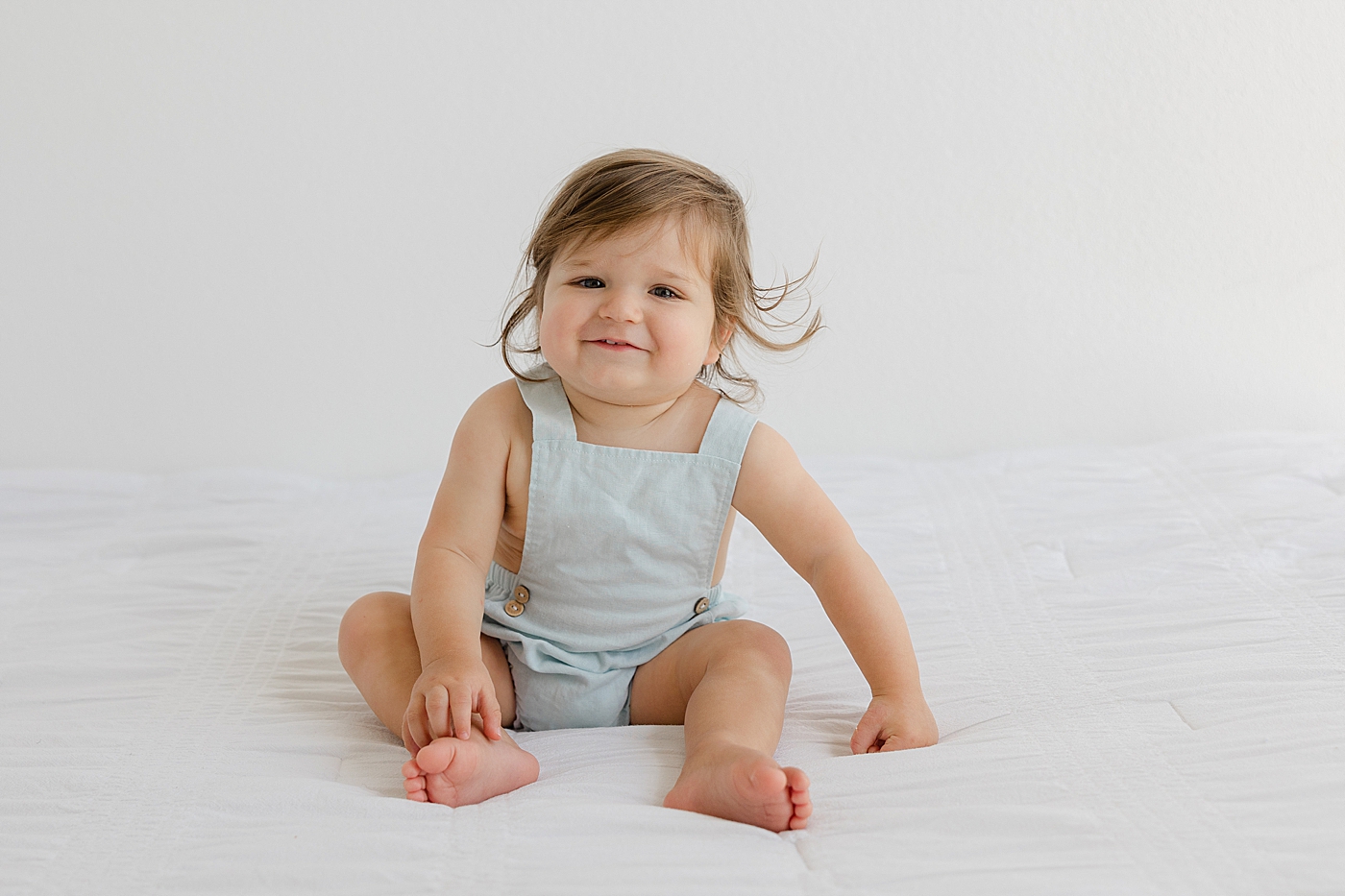 Baby girl in a light blue romper sitting on a bed | Photo by Sana Ahmed Photography