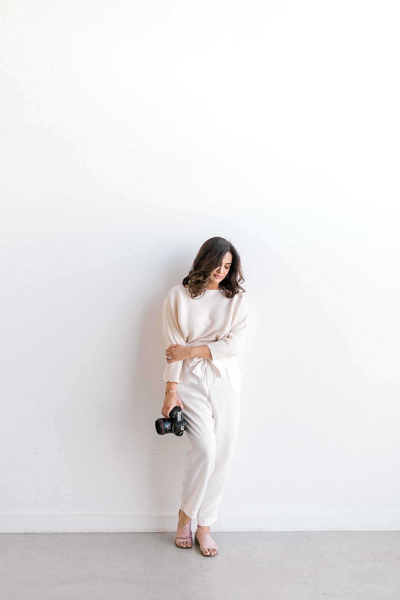 Woman in cream leaning on a wall holding her camera | Full Service Newborn Photographer Sana Ahmed 